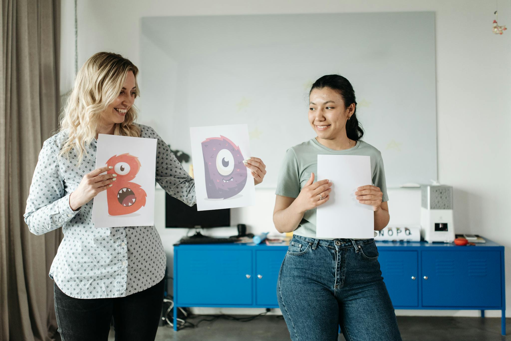 Two teachers holding fun monster drawings in a classroom setting.